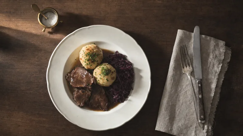 Mittagstisch im Restaurant Teller mit Rinderschmorbraten, Rotkohl und Kartoffelklößen von oben auf einem Holztisch
